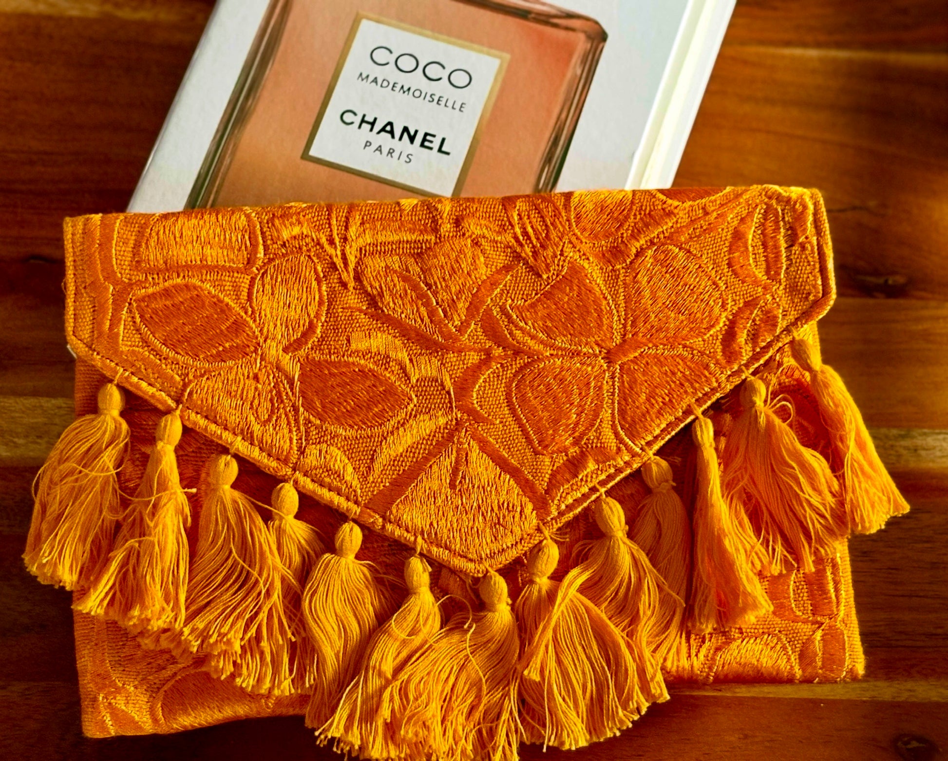 Orange textured clutch with tassels on a wooden surface with a Chanel book.