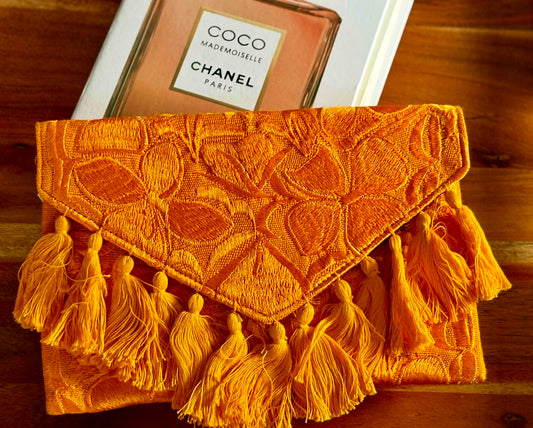 Orange textured clutch with tassels on a wooden surface with a Chanel book.