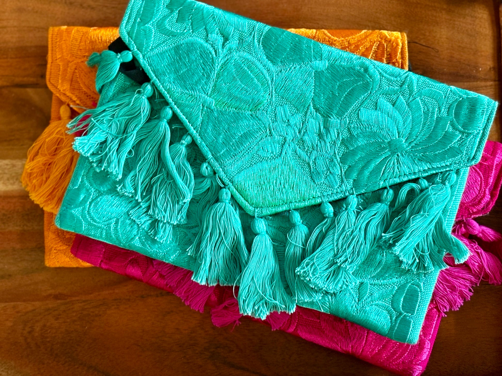 Turquoise clutch with tassels on top of pink and orange clutches on a wooden surface